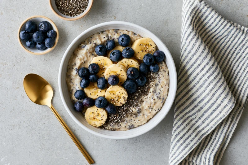 Creamy oatmeal topped with banana slices, blueberries and chia seeds in a white bowl on a gray countertop with a golden spoon and striped napkin beside it
