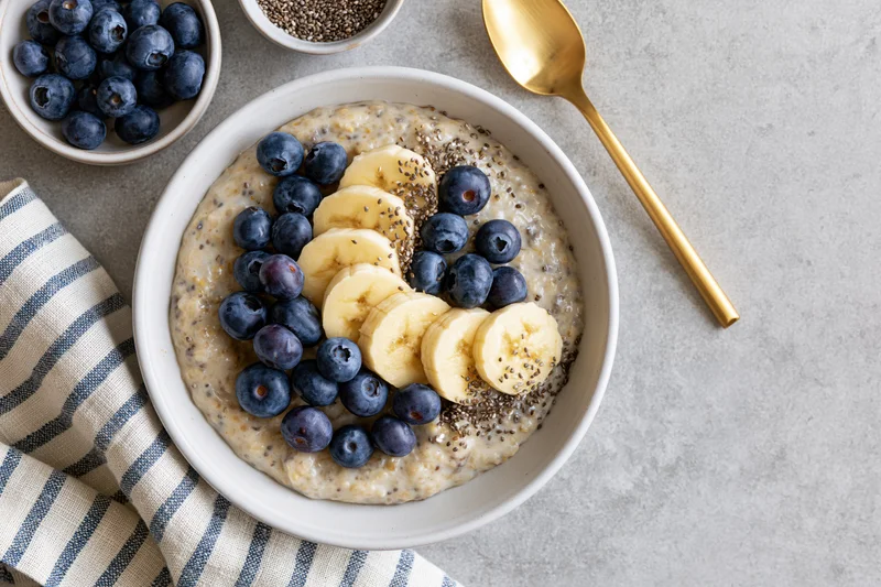 Creamy oatmeal topped with banana slices and blueberries in a white bowl on a gray surface with a striped napkin and golden spoon beside it