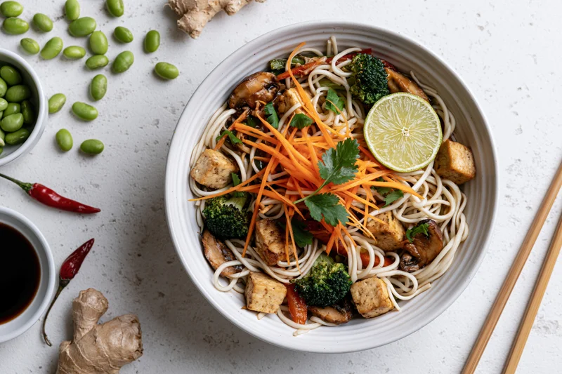 Overhead flat lay of a white ceramic bowl filled with stir-fried noodles, tofu, broccoli, carrots, lime, and cilantro, surrounded by edamame, ginger, chili, soy sauce, and chopsticks on a textured gray surface