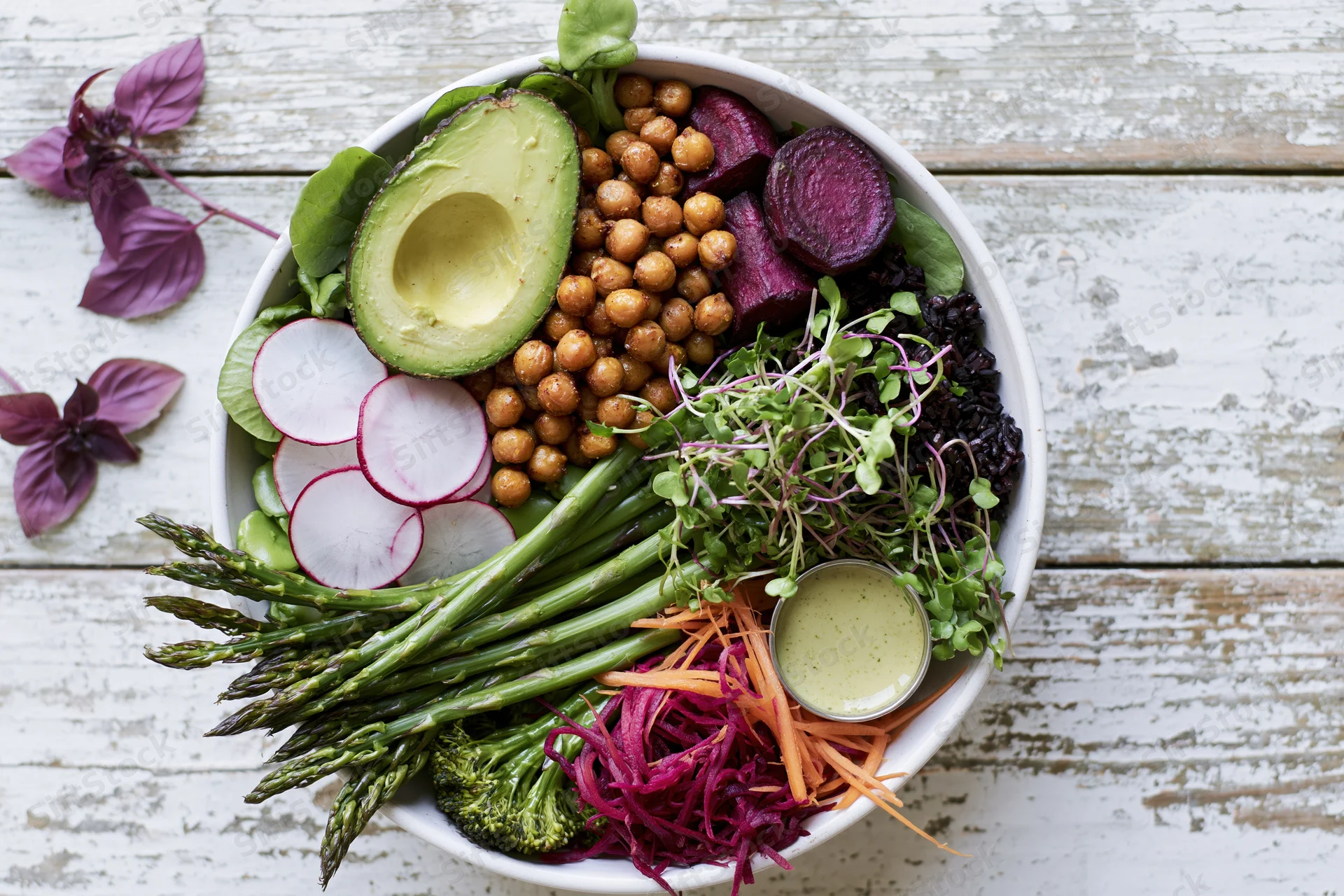 Overhead flat lay of a white ceramic buddha bowl filled with avocado, chickpeas, beetroot, radish, asparagus, broccoli, carrot, sprouts and dressing on a rustic wooden table