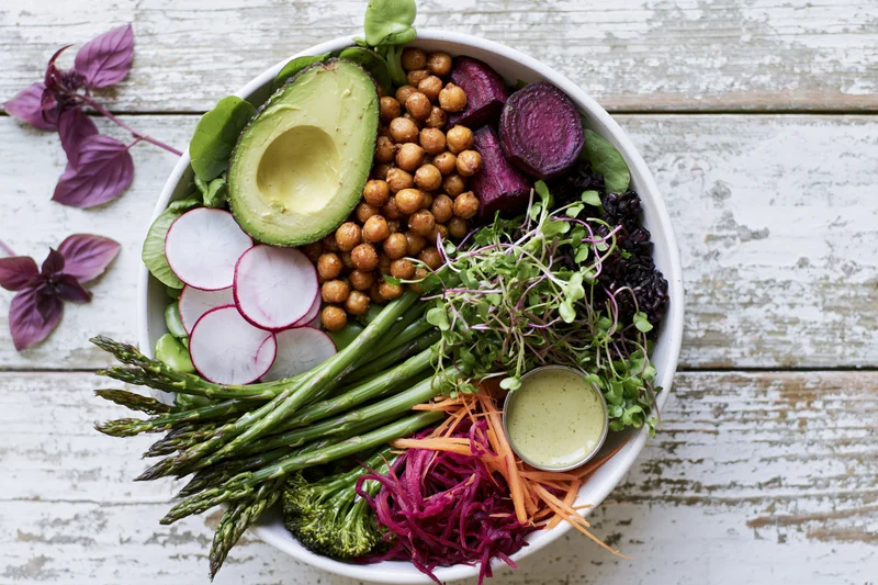 Overhead flat lay of a white ceramic buddha bowl filled with avocado, chickpeas, beetroot, radish, asparagus, broccoli, carrot, sprouts and dressing on a rustic wooden table