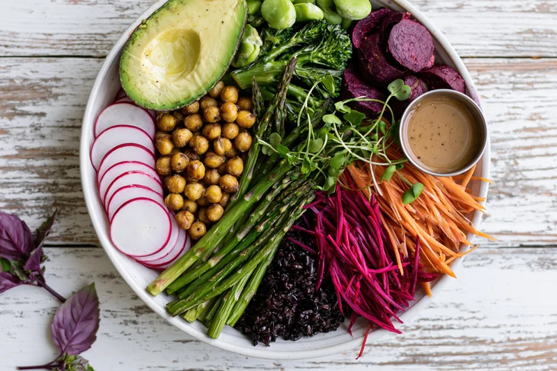 Overhead flat lay of a white ceramic buddha bowl filled with colorful vegetables, chickpeas, black rice, and dressing on a rustic wooden table