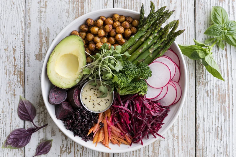 Overhead flat lay of a white ceramic buddha bowl filled with colorful vegetables, avocado, chickpeas, rice, and dressing on a rustic wooden table