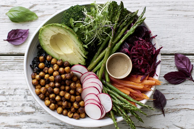 Overhead view of a white ceramic buddha bowl filled with roasted chickpeas, avocado, asparagus, radish, beetroot, carrot, broccoli, and microgreens on a rustic wooden surface