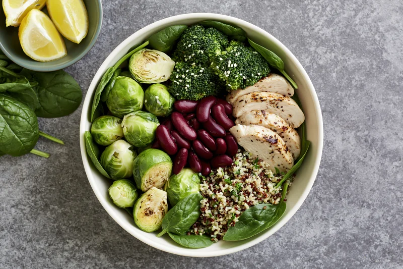 Overhead view of a white ceramic bowl filled with quinoa, broccoli, brussels sprouts, spinach, chicken, and kidney beans on a gray countertop