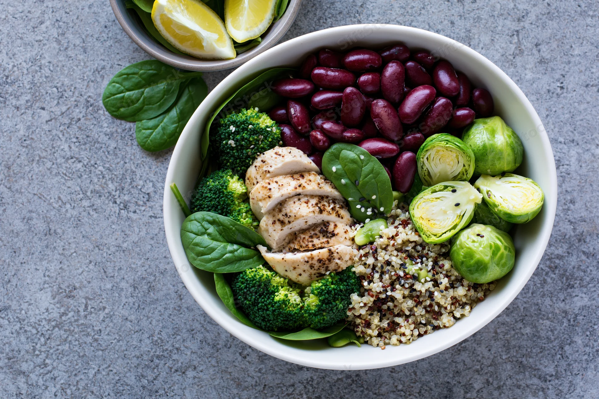 White ceramic bowl filled with quinoa, sliced chicken, broccoli, brussels sprouts, kidney beans, and spinach on gray stone surface