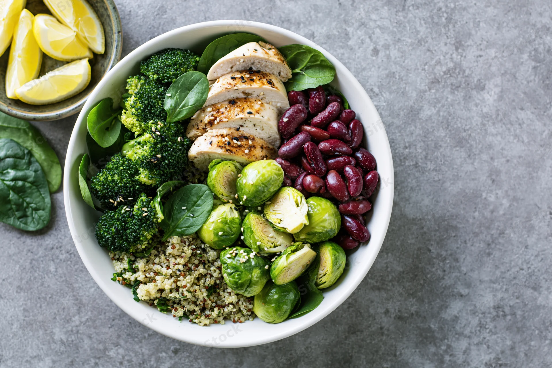 Overhead view of a white ceramic bowl filled with quinoa, sliced chicken, broccoli, brussels sprouts, kidney beans, and spinach on a gray surface with lemon wedges beside it