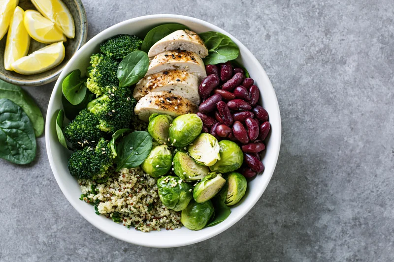 Overhead view of a white ceramic bowl filled with quinoa, sliced chicken, broccoli, brussels sprouts, kidney beans, and spinach on a gray surface with lemon wedges beside it