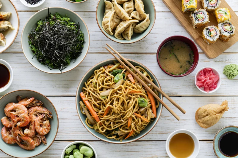 Overhead flat lay of an asian feast spread across a white wooden table with noodles, sushi, dumplings, shrimp, seaweed, miso soup, soy sauce, wasabi, pickled ginger, tempura, and chopsticks