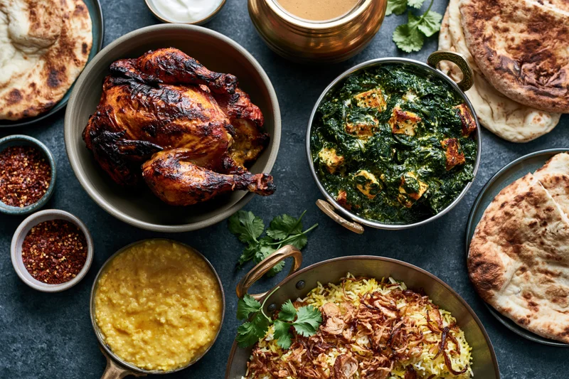 Indian feast spread with tandoori chicken, palak paneer, biryani, naan, and chutneys on dark slate surface