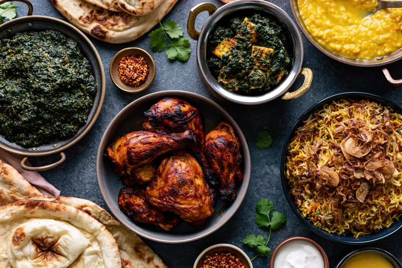 Overhead flat lay of an indian feast with tandoori chicken, biryani, palak paneer, dal, naan and cilantro on a dark slate surface