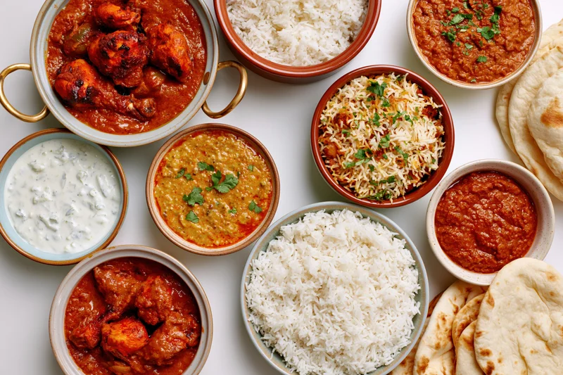 Overhead flat lay of an indian food spread with multiple bowls of curry, rice, naan and raita on a white surface