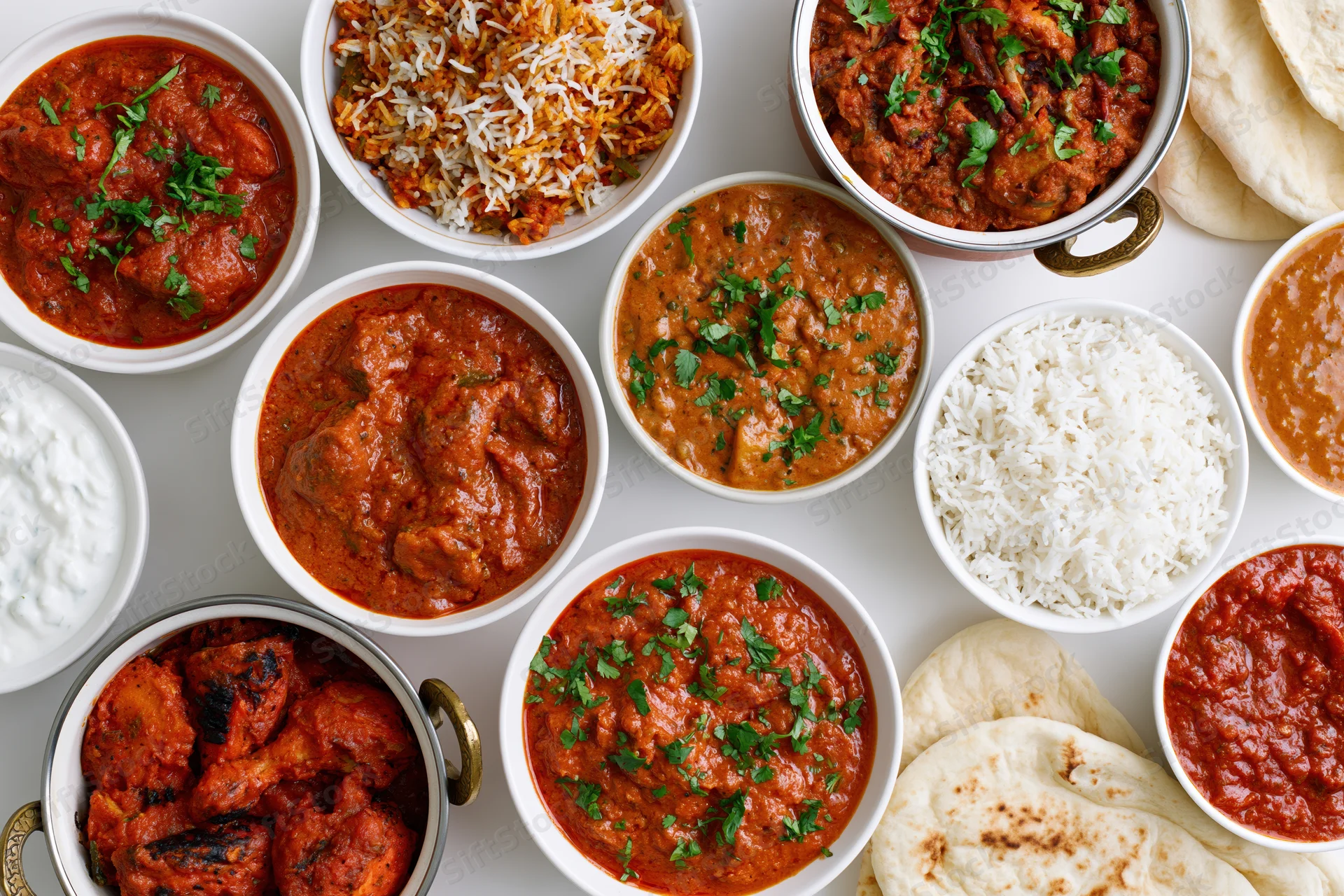 Overhead flat lay of an indian food spread with multiple bowls of curry, rice, naan, and yogurt on a white surface