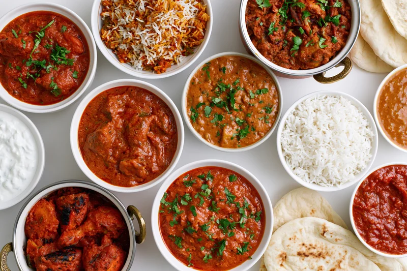 Overhead flat lay of an indian food spread with multiple bowls of curry, rice, naan, and yogurt on a white surface