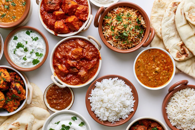 Overhead flat lay of an indian food spread with multiple bowls of curry, rice, naan, and raita arranged on a white surface