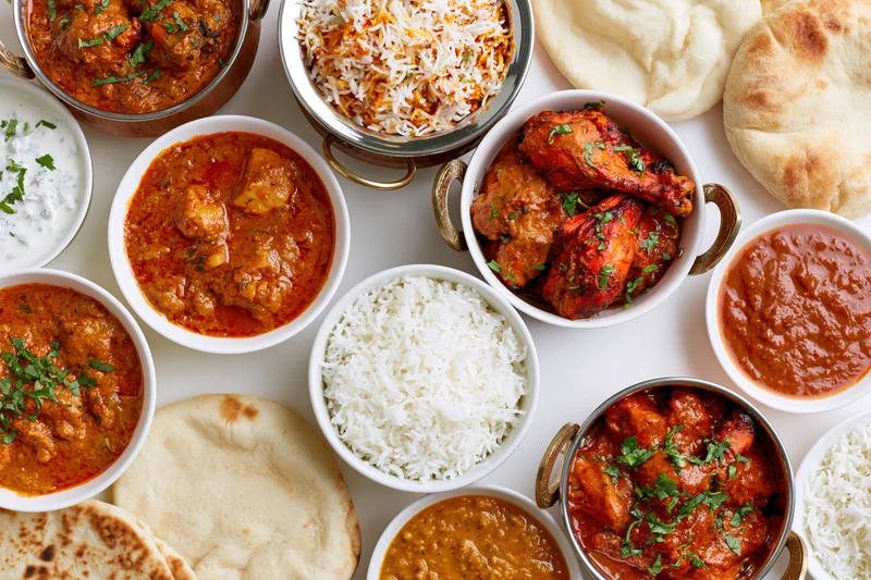 Overhead flat lay of an indian food spread with multiple bowls of curry, rice, naan and sauces arranged on a white surface