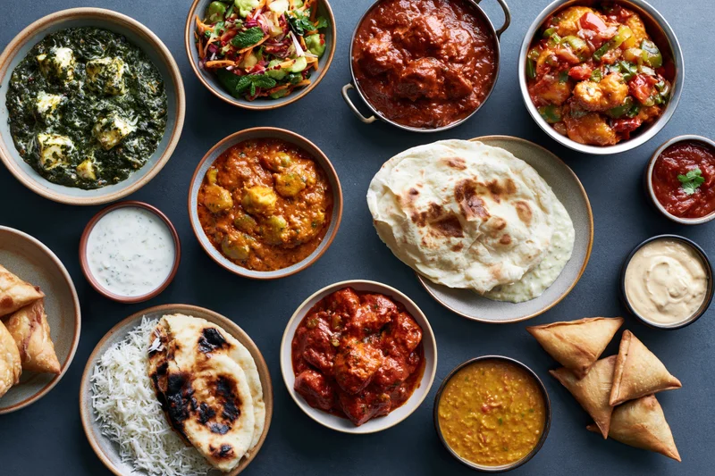 Overhead flat lay of an indian meal spread across a dark slate surface with various curries, breads, and side dishes arranged in bowls and plates