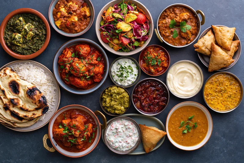 Overhead flat lay of an indian meal spread across a dark slate surface with various curries, naan, rice, samosas, and chutneys in ceramic and metal bowls