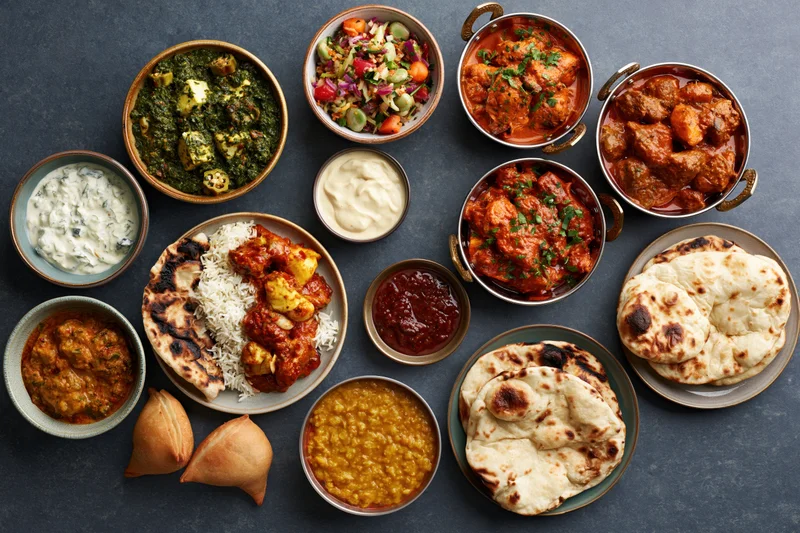 Overhead flat lay of an indian meal spread with curries, rice, naan, samosas and chutneys on a dark slate surface