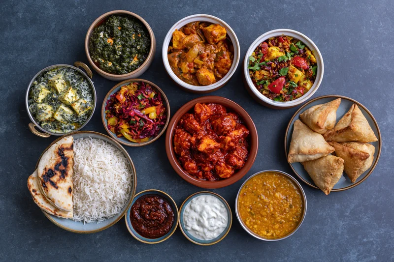 Overhead flat lay of an indian meal spread with curry, rice, naan, samosas and chutneys on dark slate surface