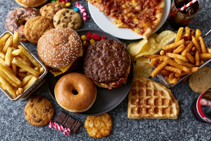 Overhead flat lay of assorted junk food spread across a dark surface including burgers, fries, pizza, donuts, waffles, cookies, soda and candy