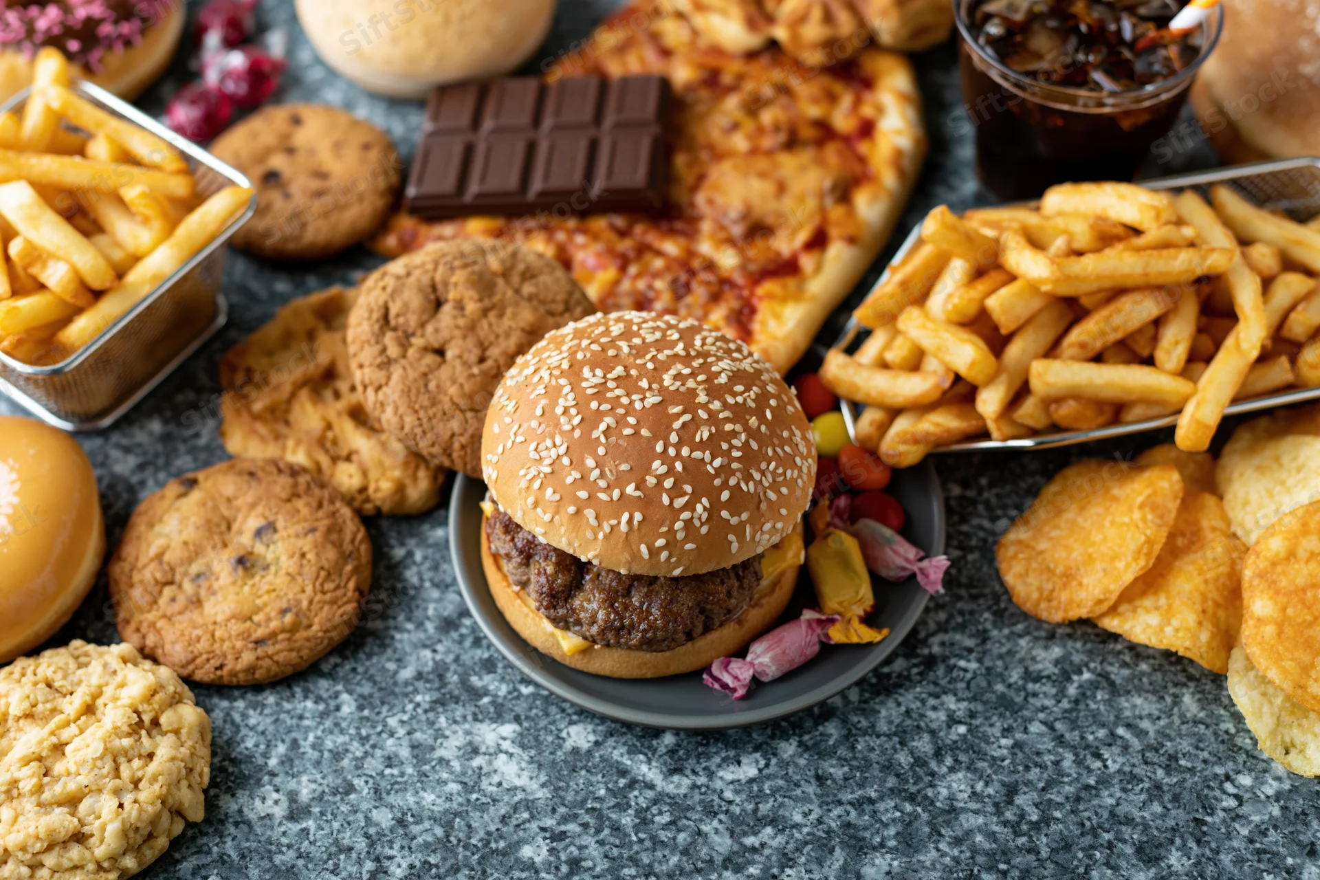 Overhead flat lay of assorted junk food including burger, fries, pizza, cookies, candy and soda spread on gray surface