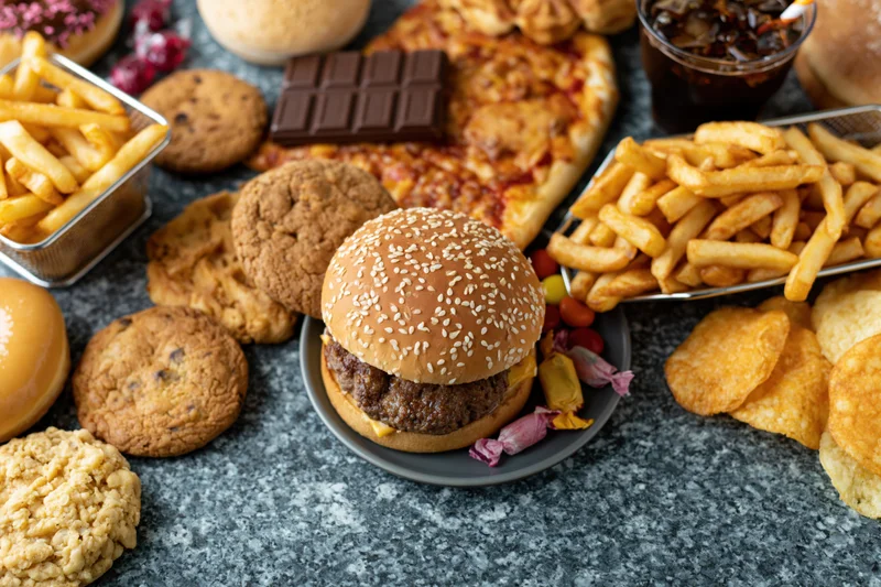 Overhead flat lay of assorted junk food including burger, fries, pizza, cookies, candy and soda spread on gray surface