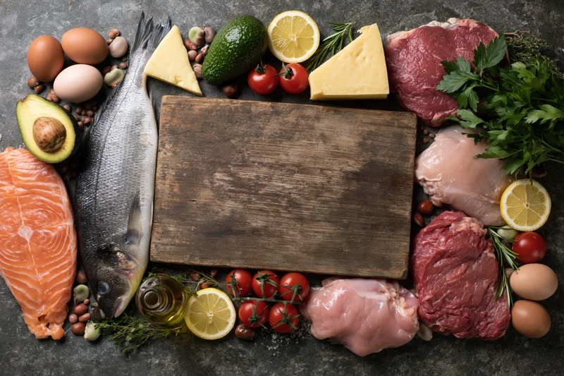 Empty wooden cutting board surrounded by raw meats, fish, eggs, vegetables and herbs on dark surface