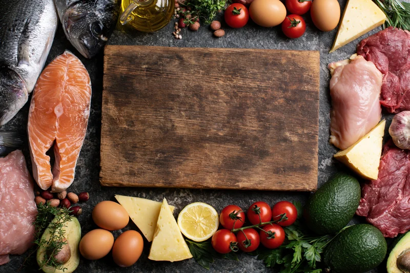 Empty wooden cutting board surrounded by raw food ingredients arranged in flat lay overhead view