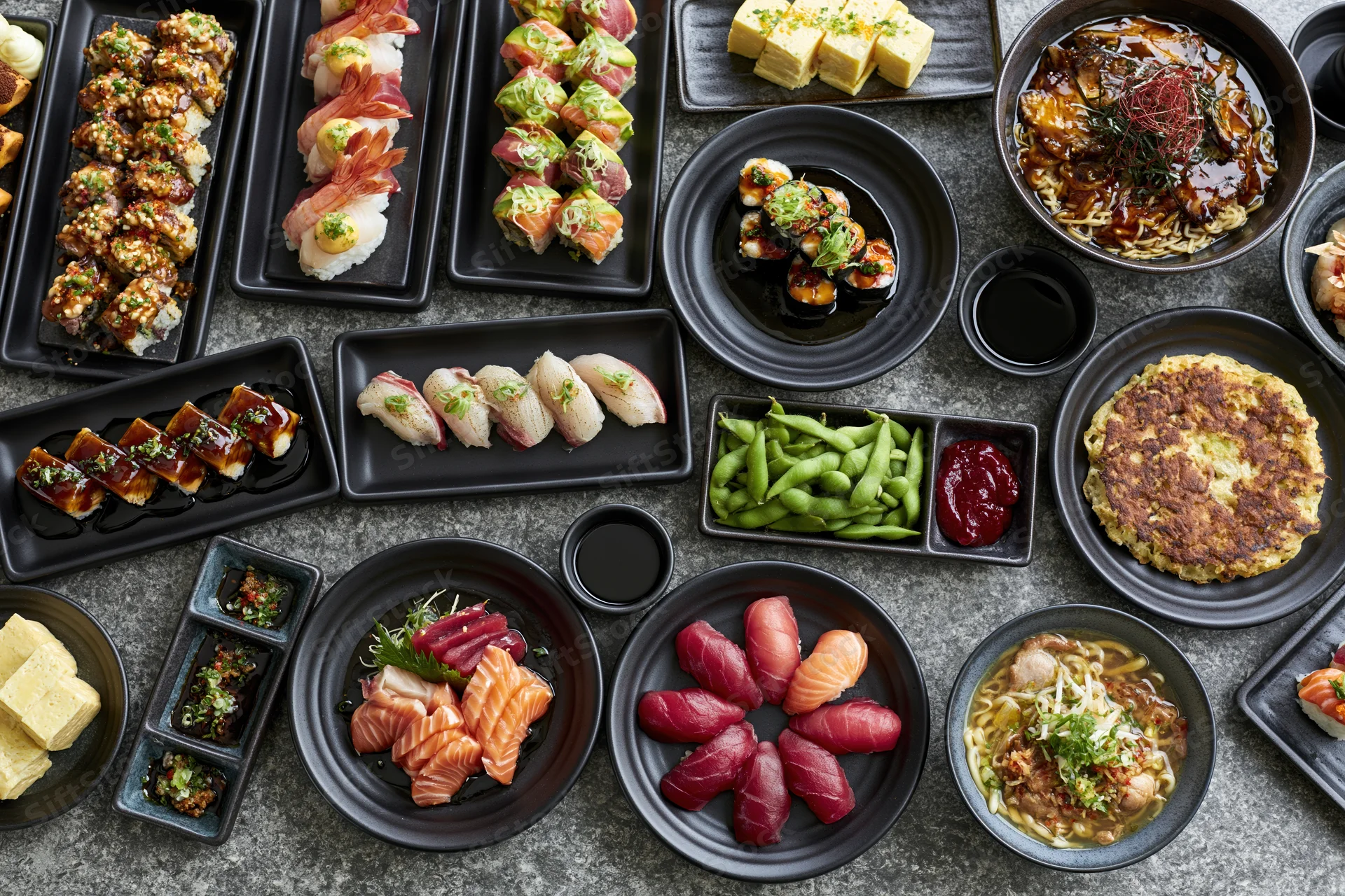 Overhead flat lay of an extensive japanese restaurant food spread on dark stone table