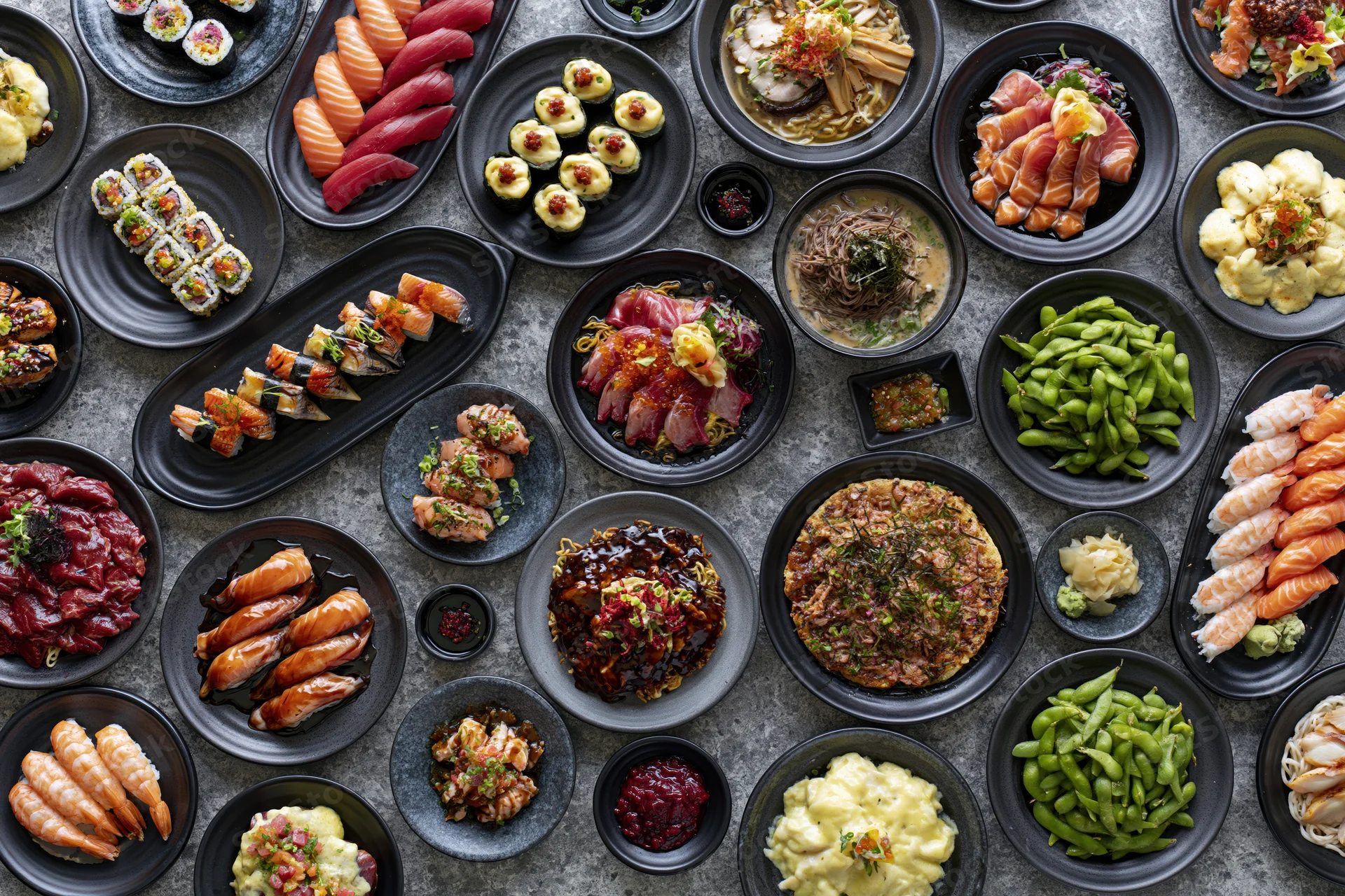 Overhead flat lay of an extensive japanese restaurant spread with multiple dishes arranged on black plates