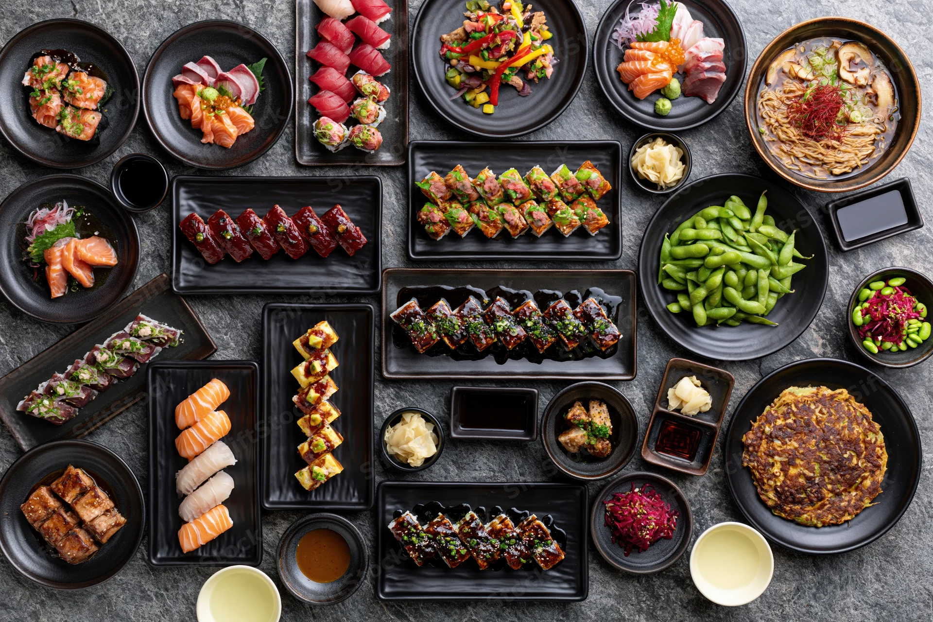 Overhead flat lay of an extensive japanese restaurant spread with sushi, sashimi, edamame and various dishes arranged on black plates on a stone surface