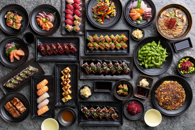 Overhead flat lay of an extensive japanese restaurant spread with sushi, sashimi, edamame and various dishes arranged on black plates on a stone surface