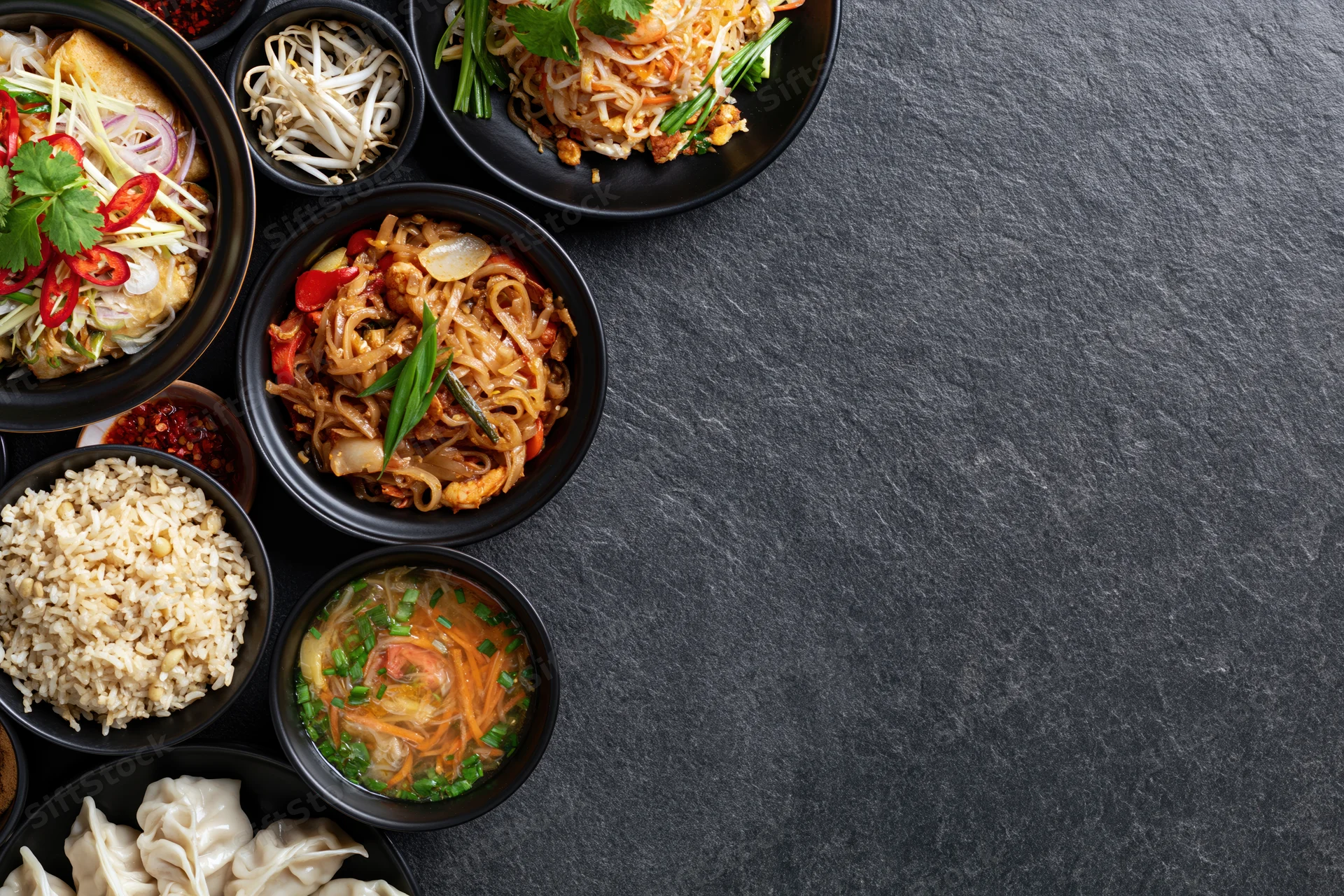 Assorted asian dishes arranged in black bowls on dark slate surface with copyspace