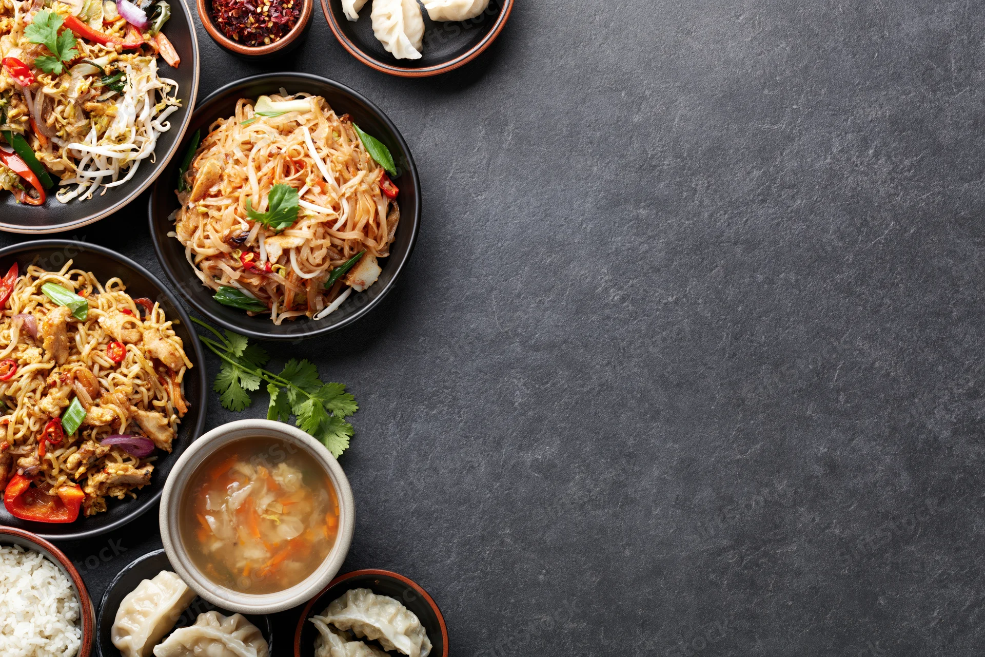 Assorted asian dishes arranged along the left side of a dark stone table in overhead flat lay view