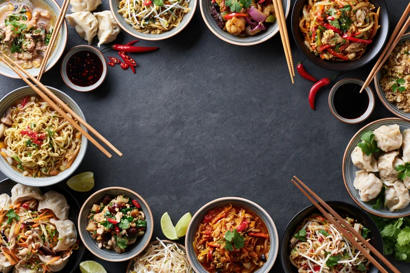 Assorted asian dishes arranged in a border around a dark table with chopsticks and garnishes