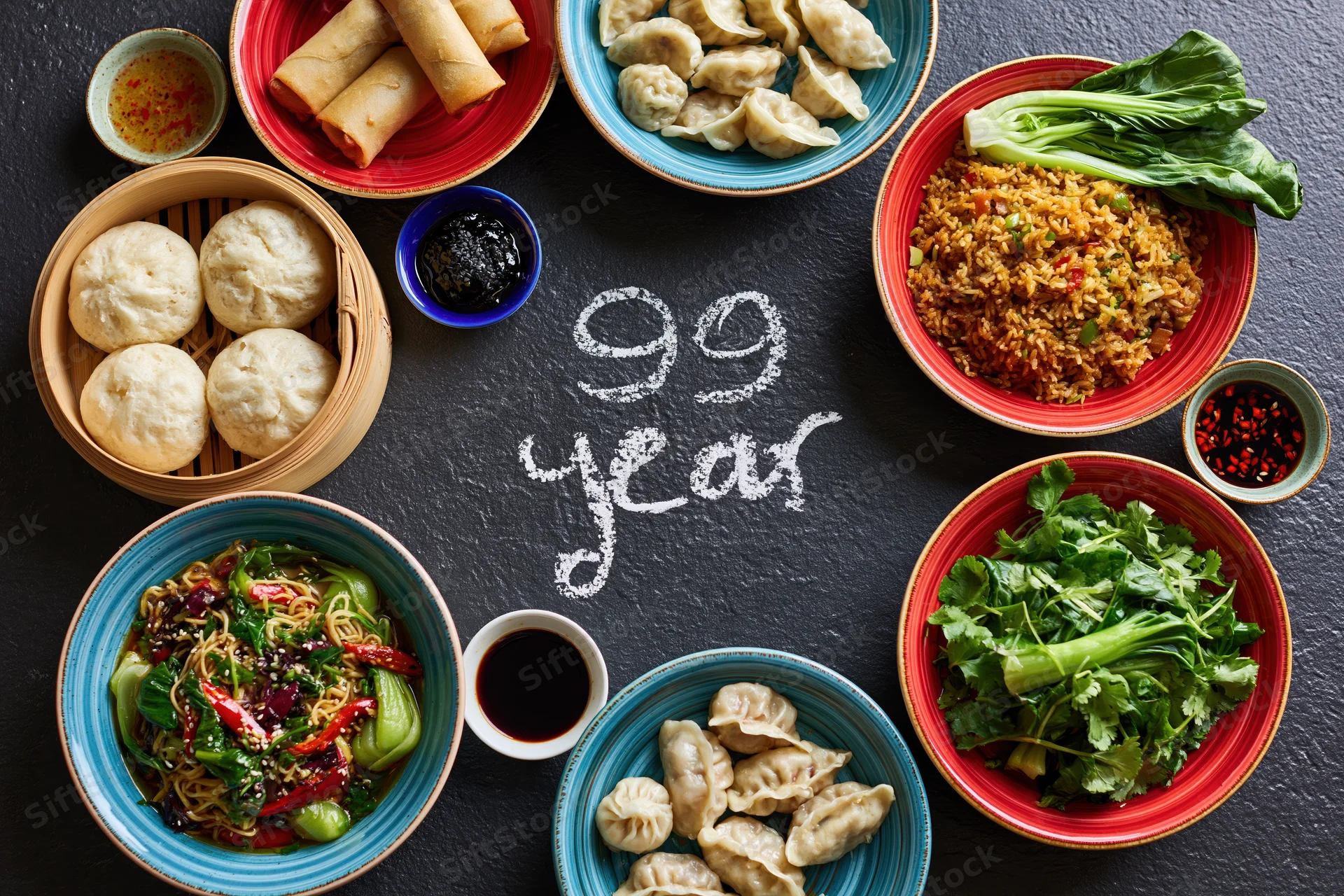Assorted chinese dishes arranged in a circle around chalk-written 99 year on dark slate surface