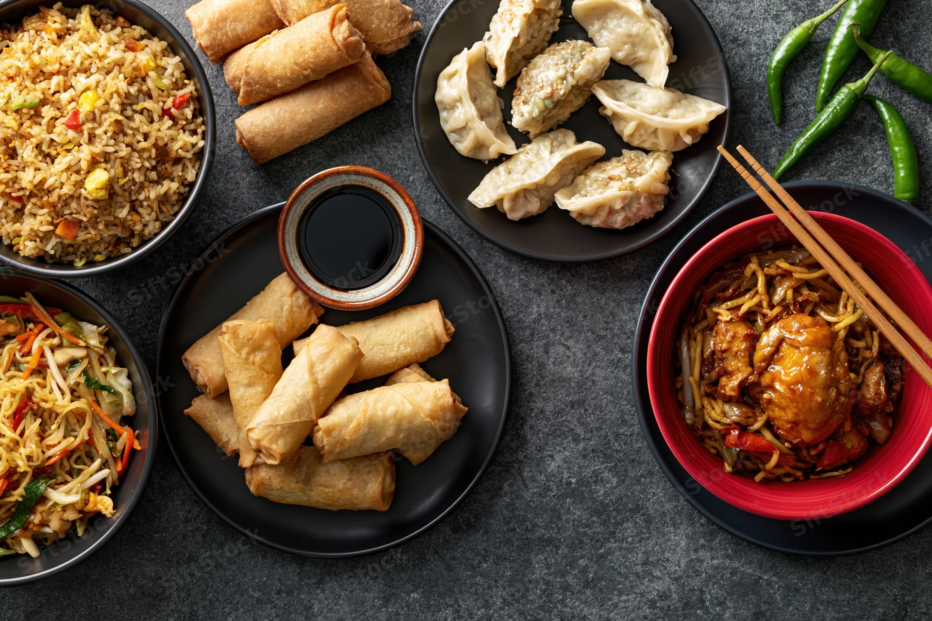Overhead flat lay of assorted chinese dishes arranged on dark stone surface with chopsticks and chili peppers