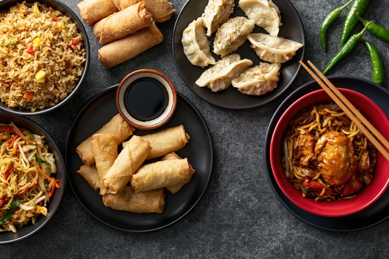 Overhead flat lay of assorted chinese dishes arranged on dark stone surface with chopsticks and chili peppers
