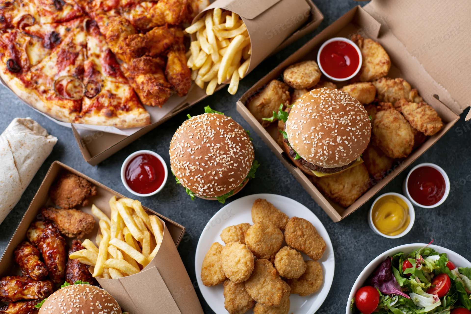 Overhead flat lay of assorted fast food including burgers, pizza, fries, chicken wings, nuggets and salad on dark surface