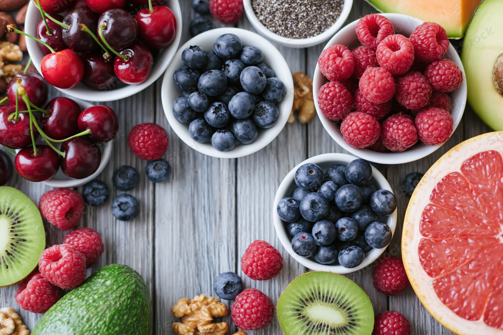 Overhead flat lay of assorted fresh berries fruits nuts and chia in white bowls on wooden table