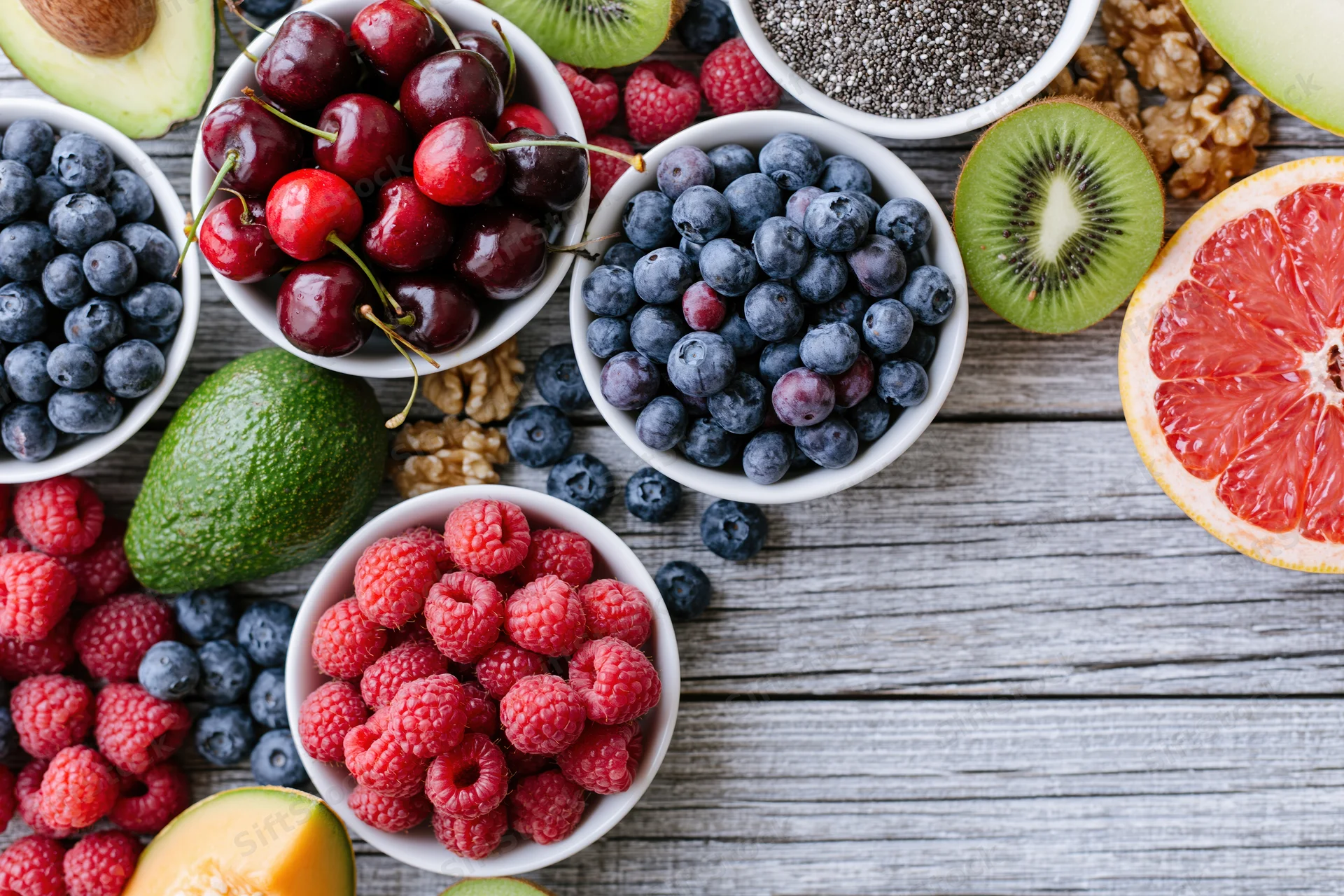Overhead flat lay of assorted fresh berries fruits nuts and chia in white bowls on wooden table
