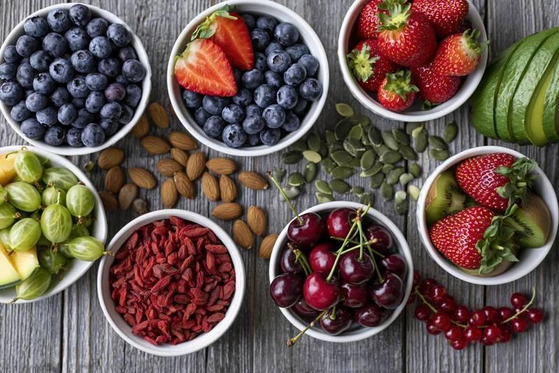 Overhead flat lay of assorted fresh fruits berries nuts and seeds in white bowls on wooden surface