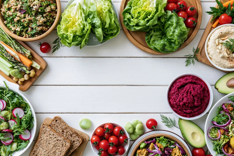 Overhead flat lay of assorted healthy vegetarian dishes and snacks arranged on a white wooden table with copyspace in center