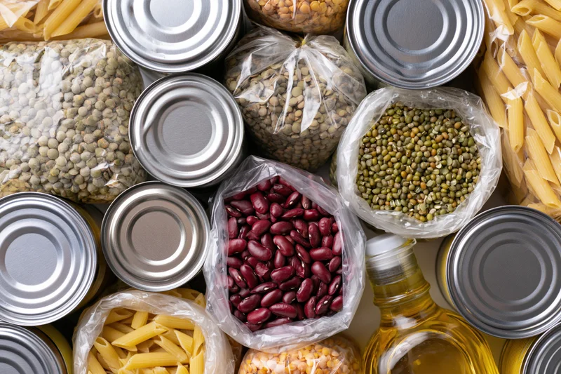 Overhead flat lay of assorted pantry staples including canned goods, dry beans, pasta, and cooking oil arranged on a surface