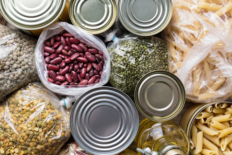 Overhead flat lay of pantry staples including canned goods, beans, pasta, and legumes on surface