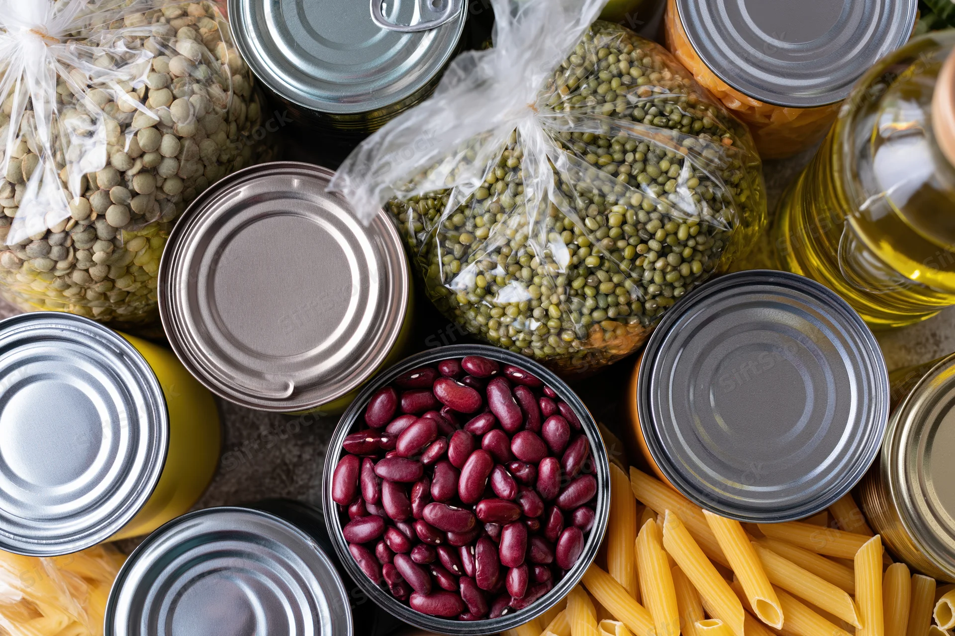 Overhead flat lay of canned beans, pasta, oil, and legumes arranged on a dark surface
