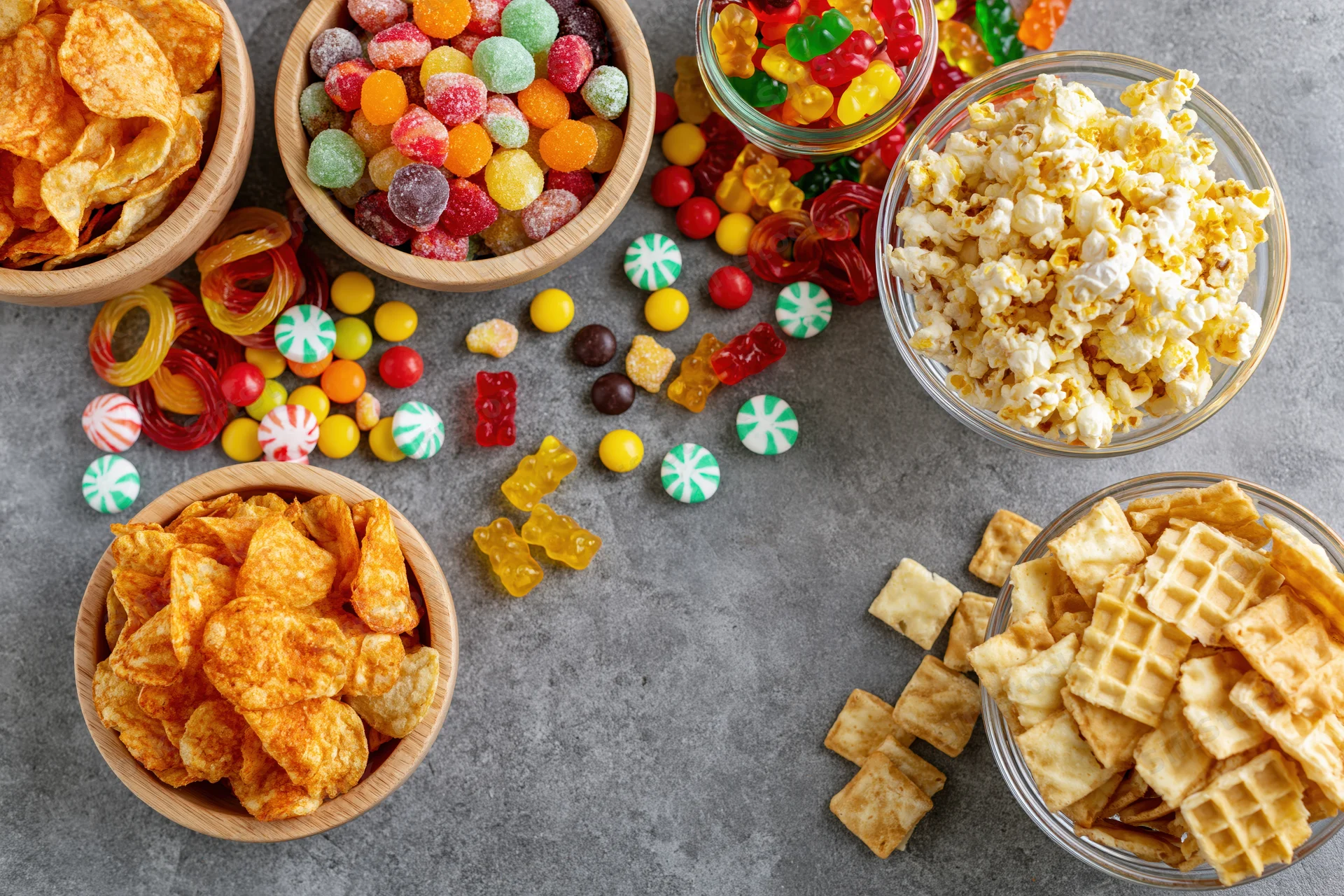 Assorted snacks and candies arranged in bowls and scattered on a gray surface in overhead flat lay view