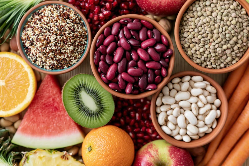 Overhead flat lay of assorted whole foods densely arranged on wooden surface with bowls of beans, quinoa, lentils, fruits and vegetables