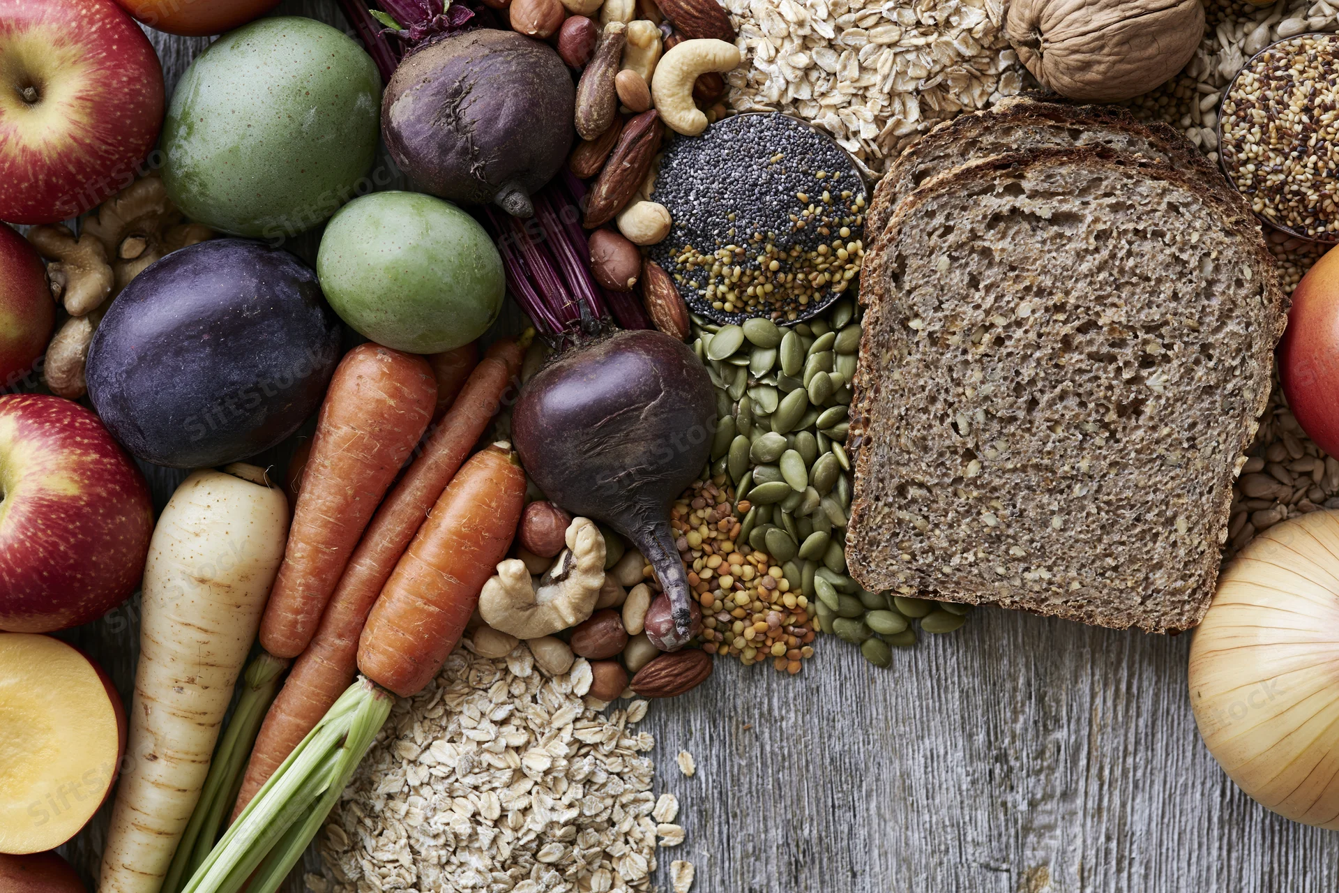 Overhead flat lay of whole foods including vegetables, fruits, grains, nuts, and seeds arranged on wooden surface